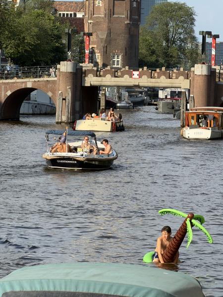 This vibrant photo captures a lively summer afternoon on the canals of Oudeschans, Uilenburg, Amsterdam, with several boats navigating the water and a person playfully floating on an inflatable palm tree
